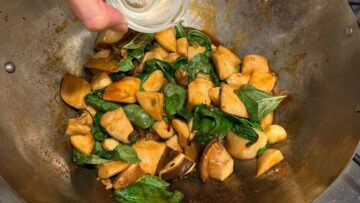 Fresh mushrooms and basil being stir-fried in a wok, as a hand pours sauce from a small glass bowl—perfect for creating a vegetarian three cup chicken-inspired dish.