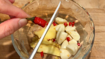 A hand slicing a red chili pepper over a glass bowl with garlic and ginger on a wooden surface.