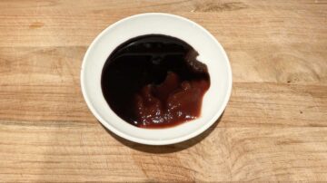 A small white dish with partial ingredients for the three cup sauce: soy sauce, vegetarian oyster sauce, ketchup, and sugar, on a wooden surface.