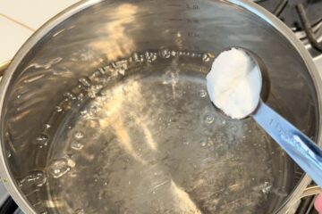 A tablespoon of sugar being added to a pot of boiling water on a stove.