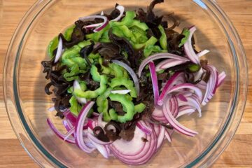 Sliced bitter melon, red onion, and woodear mushrooms in a glass bowl on a wooden surface.
