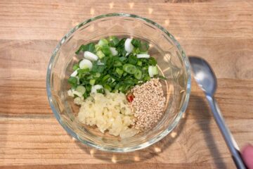 Glass bowl with chopped green onions, pressed garlic, and sesame seeds on a wooden surface.