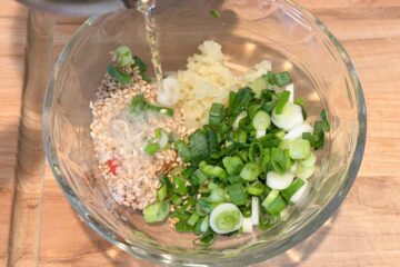 Hot oil being poured on sesame seeds in a glass bowl with chopped green onions, crushed garlic, sesame seeds, and chopped red chili peppers.