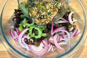 Sliced bitter melon, red onion, and woodear mushrooms in a glass bowl, with dressing being poured from a smaller glass bowl.