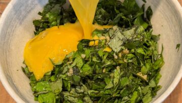 Chopped basil leaves in a bowl, with beaten eggs being poured from the top.