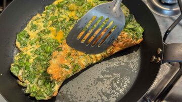 A basil omelette is being folded with a spatula in a frying pan.
