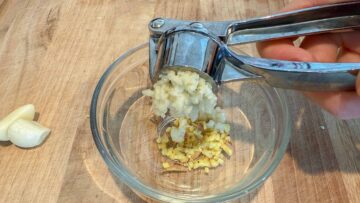 A garlic press squeezes garlic into a glass bowl with chopped ginger on a wooden surface.