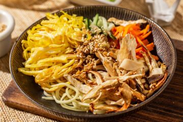 Close up of a bowl of cold sesame noodles topped with shredded king oyster mushrooms, carrots, cucumbers, eggs, sauce, and sprinkled with white sesame seeds.