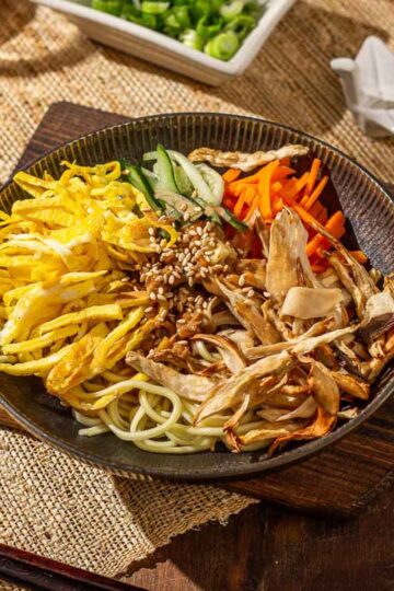 A bowl of cold sesame noodles topped with shredded king oyster mushrooms, egg, cucumber and carrot strips, and sprinkled with some white sesame seeds.