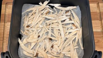 Shredded pieces of mushroom spread on parchment paper inside an air fryer basket on a wooden surface, perfect as a topping for cold sesame noodles.