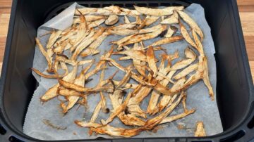 Thin strips of dried mushrooms on parchment paper in an air fryer basket, perfect as a crunchy topping for cold sesame noodles.