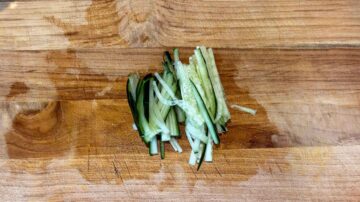 Thinly sliced cucumber strips arranged on a wooden cutting board, perfect as a fresh topping for cold sesame noodles.