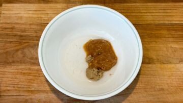 A white bowl with a brown paste and sauce, perfect for tossing with cold sesame noodles, sits on a wooden surface.