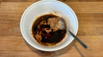 A white bowl of cold sesame noodles with brown sauce and a spoon, sitting on a wooden surface.