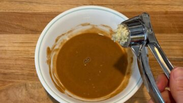 A garlic press over a bowl of brown sauce on a wooden surface, ready to add bold flavor to cold sesame noodles.