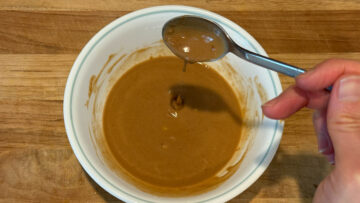 A hand holding a spoon above a bowl of creamy brown sauce for cold sesame noodles on a wooden surface.