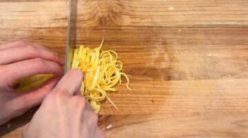 A person slicing thin yellow egg strips on a wooden cutting board with a knife, preparing toppings for cold sesame noodles.