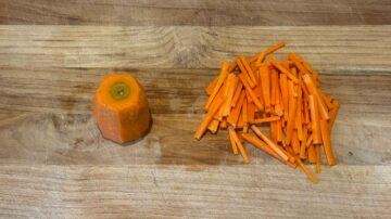 A peeled carrot and a pile of julienned carrot sticks on a wooden cutting board, perfect for adding crunch and color to cold sesame noodles.