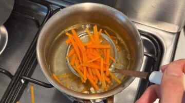 A hand adds julienned carrots to boiling water in a pot on a stovetop, preparing ingredients for delicious cold sesame noodles.
