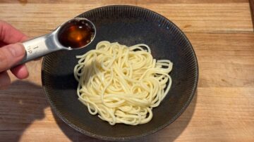 A hand pours sauce from a measuring spoon onto cold sesame noodles in a dark bowl on a wooden surface.