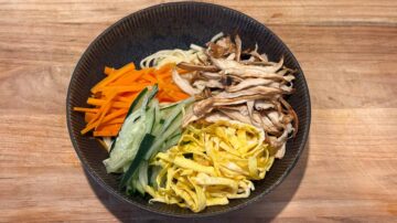 A bowl of cold sesame noodles topped with sliced carrots, cucumbers, mushrooms, and thin strips of egg sits on a wooden table.