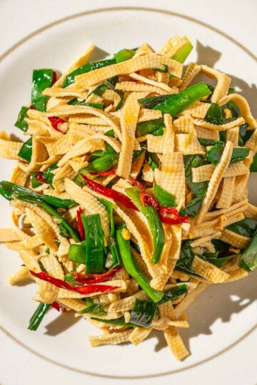 Overhead shot of a plate of Chinese Garlic Chives with Dried Tofu Noodles (and some dried chili peppers), served on a round white dish placed on a wooden surface.