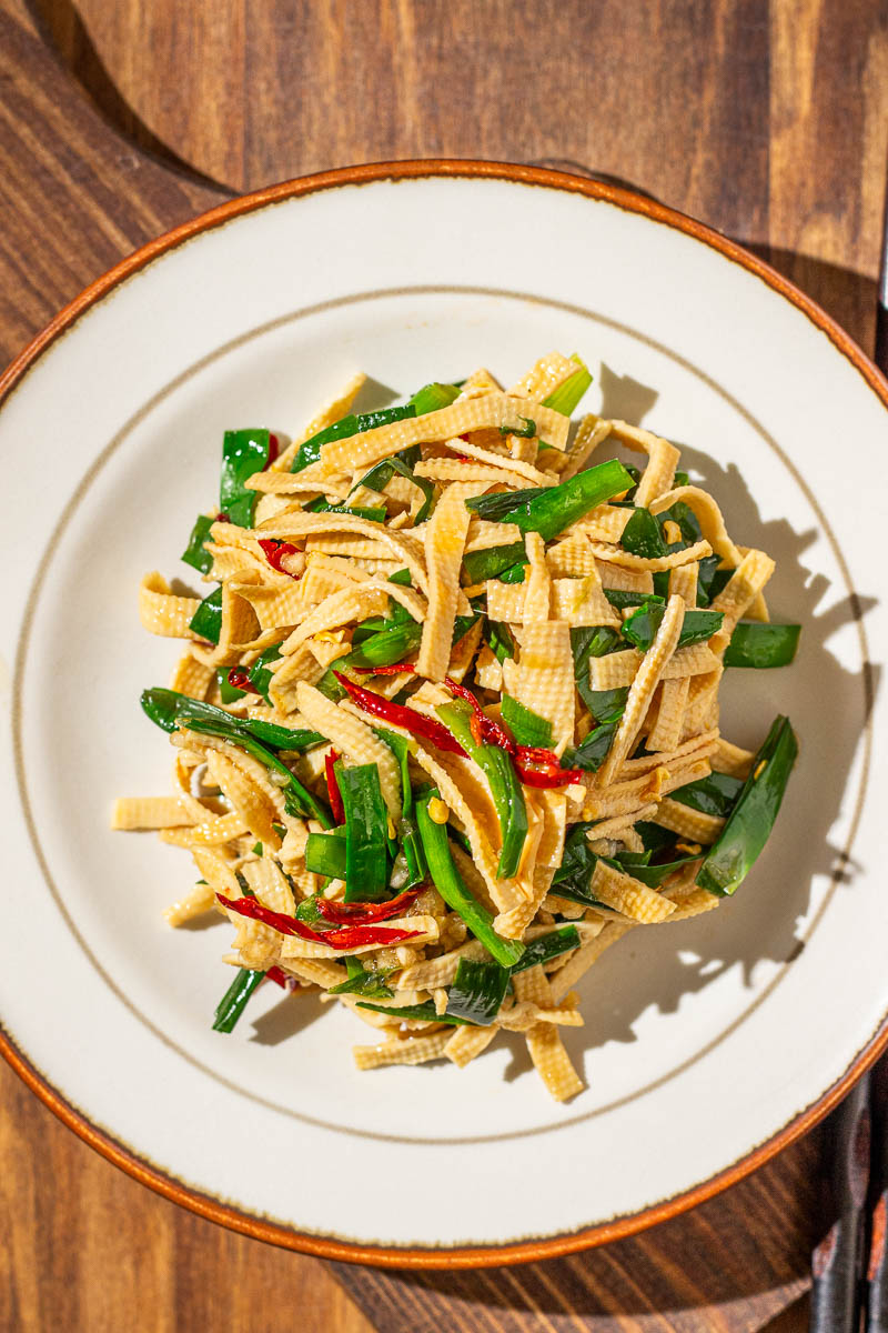 Overhead shot of a plate of Chinese Garlic Chives with Dried Tofu Noodles (and some dried chili peppers), served on a round white dish placed on a wooden surface.