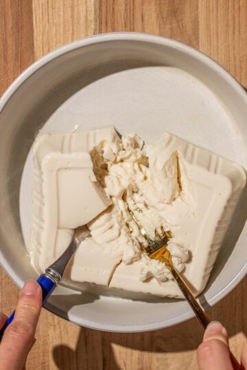 Hands mashing silken tofu in a bowl with two forks, on a wooden surface.