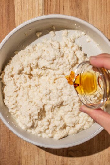 A hand pours sesame oil into a bowl of mashed silken tofu on a wooden surface.