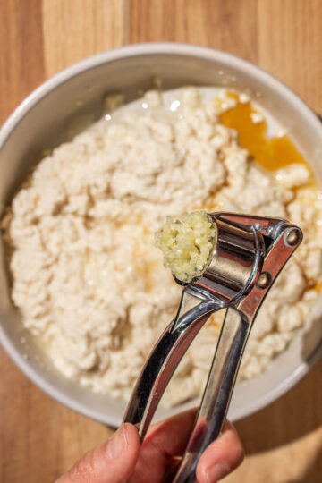 A hand holding a garlic press over a bowl of mashed silken tofu on a wooden surface.