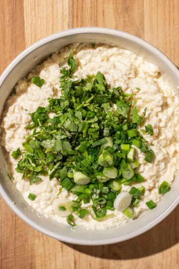 A bowl of mashed silken tofu topped with chopped green onions and cilantro on a wooden surface.