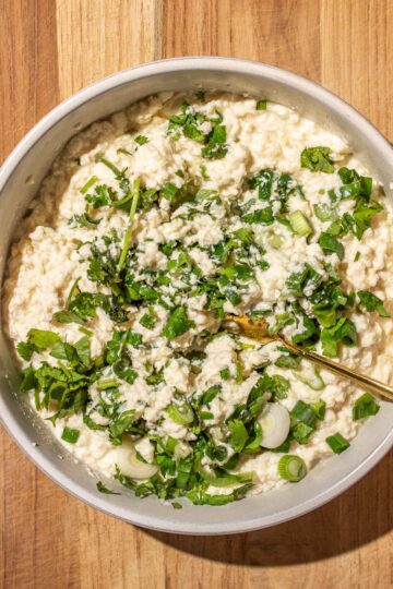 A bowl of mashed silken tofu mixed with chopped green onions and cilantro on a wooden surface.