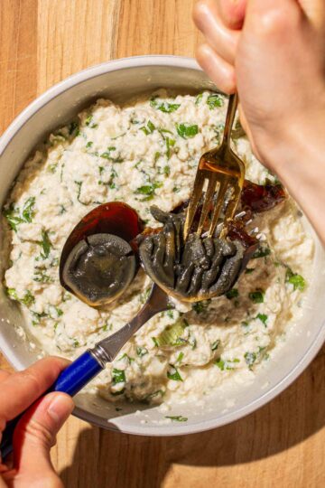 Hands using two forks to mash century eggs in a bowl of mashed silken tofu with green onions.