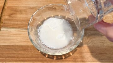 A hand pours water into a glass bowl with glutinous rice flour on a wooden surface.