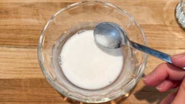 A hand mixing water and glutinous rice flour into a slurry in a small glass bowl on a wooden surface.