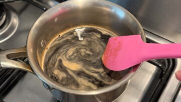 A pink spatula stirs the soy sauce and water mixture in a stainless steel pot on a stove.