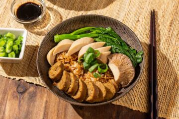 A bowl of clay pot rice topped with gai lan, slices of king oyster mushrooms and vegan sausage, and green onions, with chopsticks and sauces on the side.