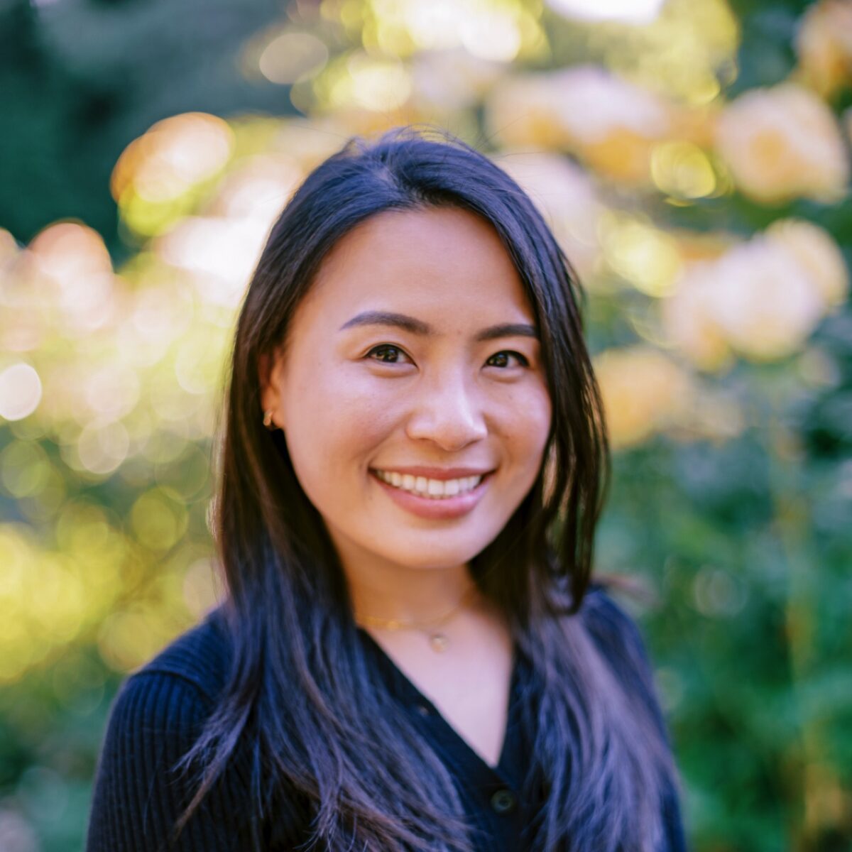 Woman with long dark hair smiling outdoors, among blurred greenery and yellow flowers in the background.