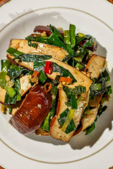 An overhead shot of a plate of Chinese Garlic Chives Bean Curd Stir Fry, with bits of chopped red chili peppers and minced garlic on top, served in a white dish with brown rim on a wooden table.