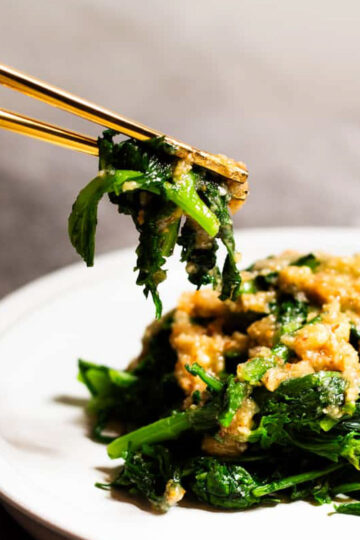 Side view of a pair of chopsticks holding chrysanthemum greens (tong hao) with mixed nut sauce, above a plate of the same.