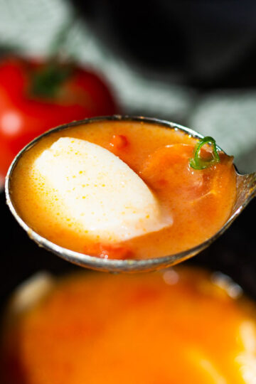 A spoonful of Tomato Rice Cake Soup—with a piece of rice cake, tomato, and green onions—above a black bowl, fresh tomatoes in the background.