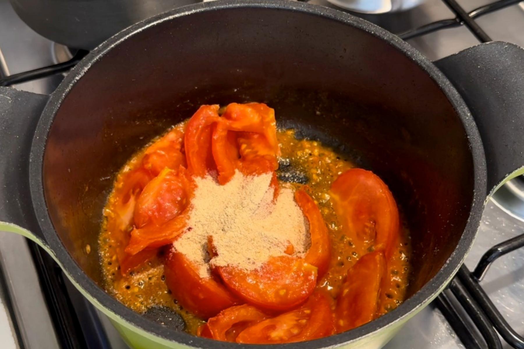 Tomato wedges and nutritional yeast cooking in a black pot on the stovetop, creating the flavorful base for Tomato Rice Cake Soup.