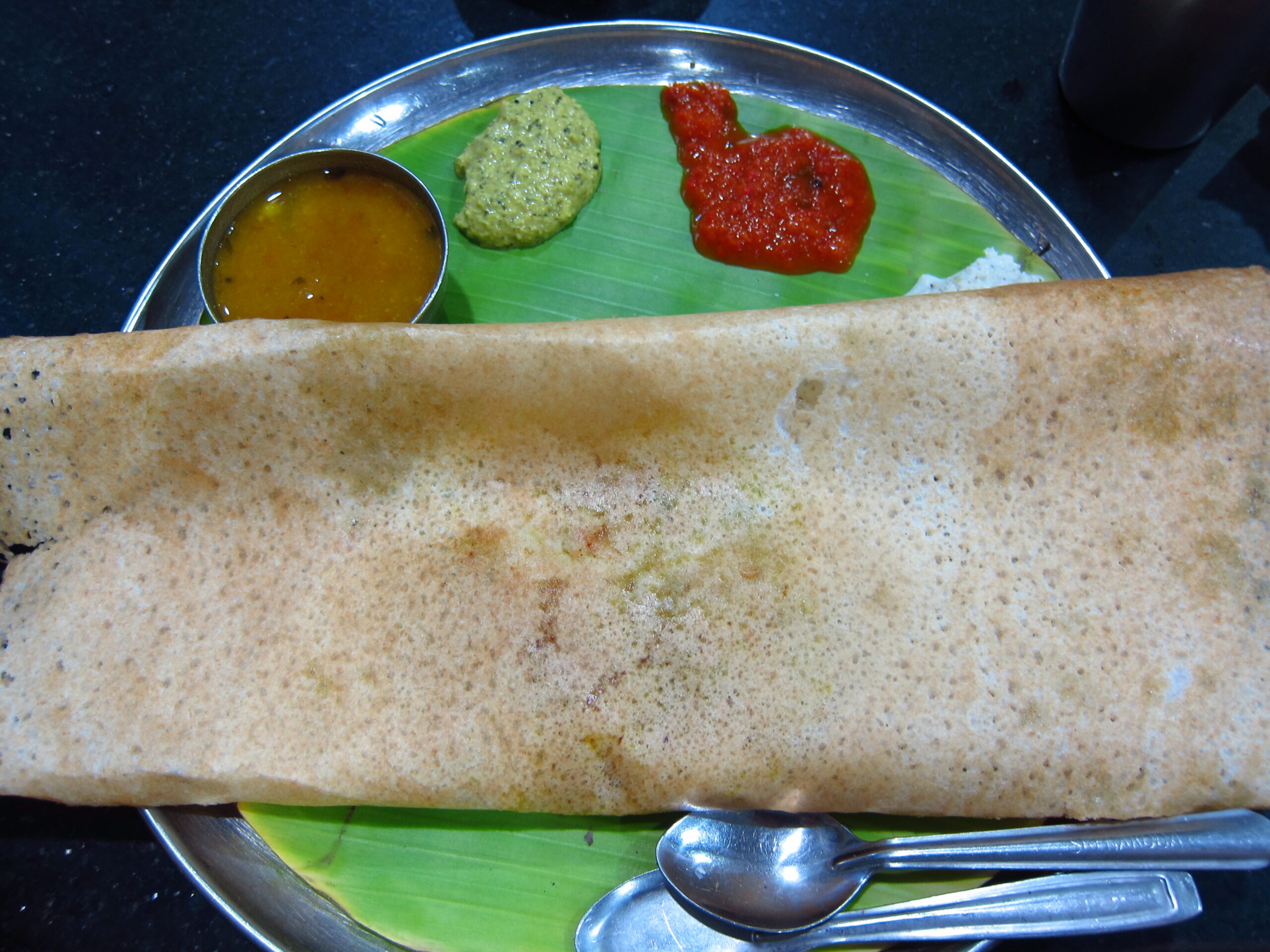 A dosa on a banana leaf with sambar, green chutney, red chutney, and cutlery on a metal plate.