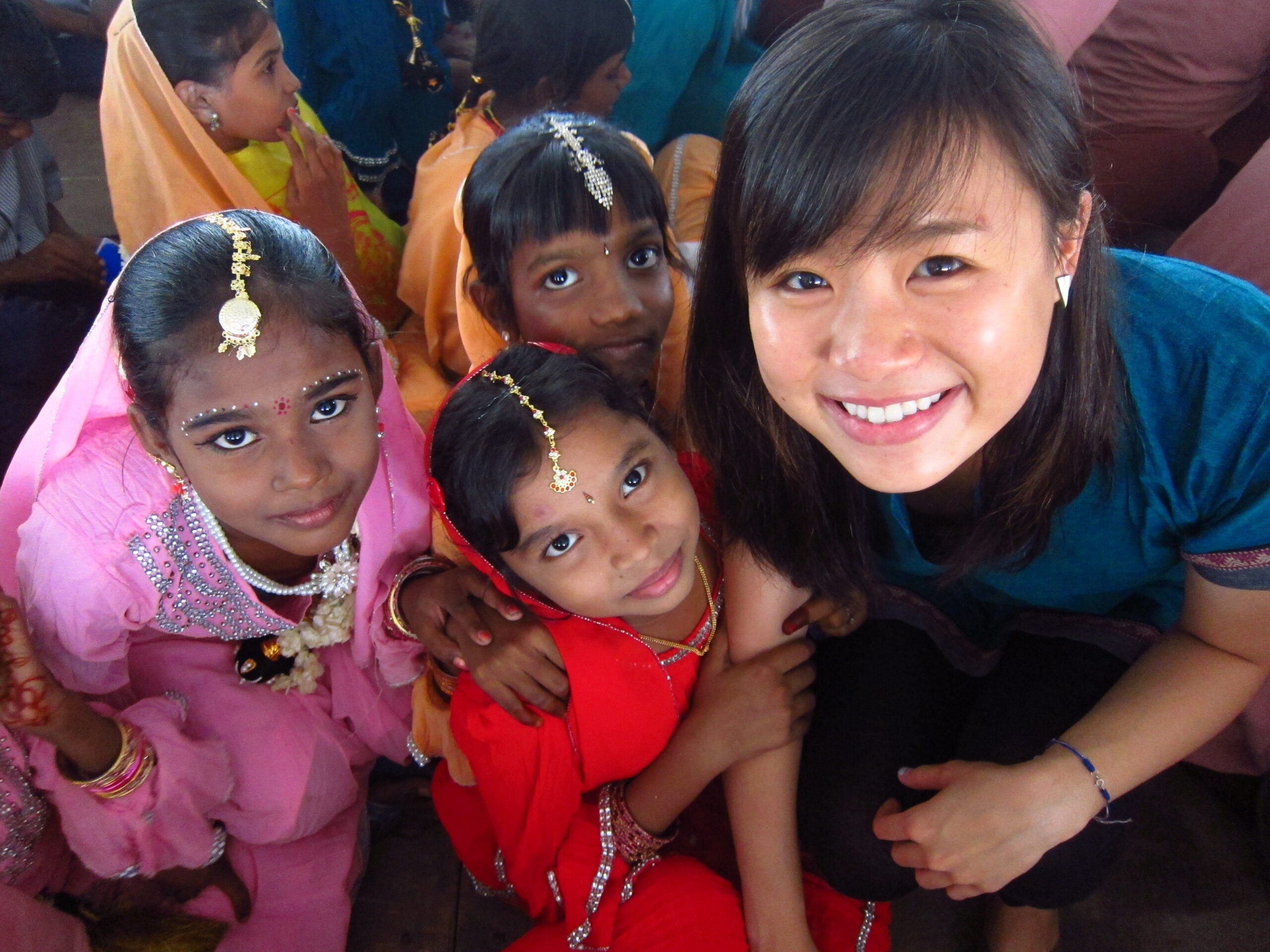 Four young girls in colorful traditional clothing and jewelry smile at the camera with a woman in a blue shirt.