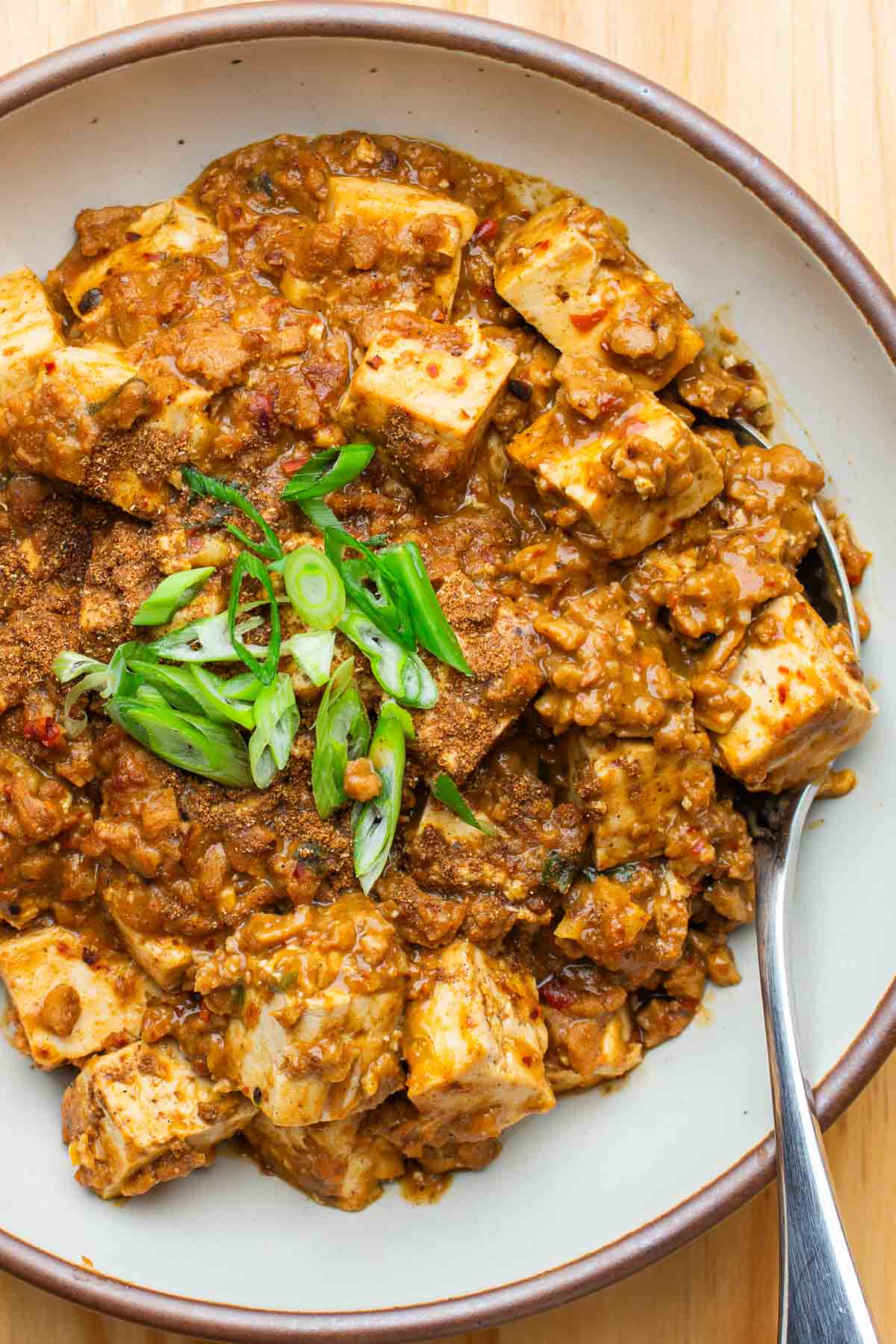 A bowl of vegan mapo tofu topped with sliced green onions, with a spoon on the side.