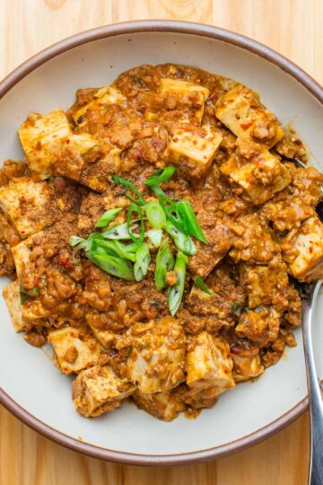 A bowl of vegan mapo tofu, topped with sliced green onions and with a spoon on the side, on a wooden table.