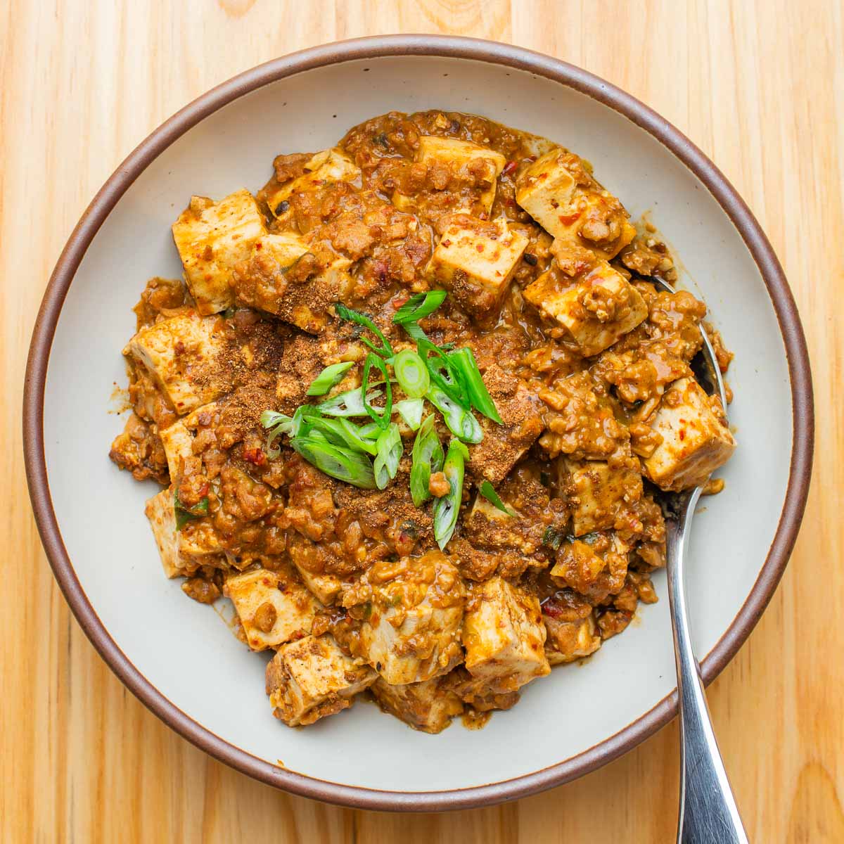 A bowl of vegan mapo tofu, topped with sliced green onions and with a spoon on the side, on a wooden table.