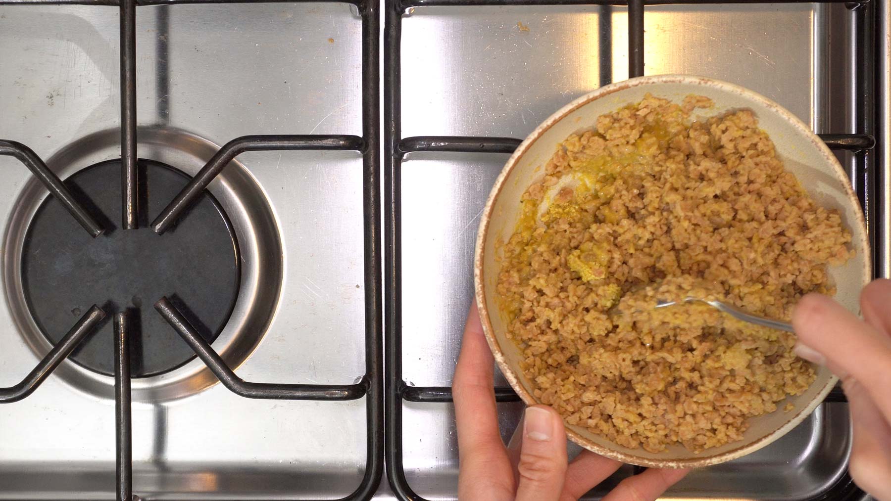 Hands mixing Textured Vegetable Protein (TVP), nutritional yeast, and marinating liquid with a spoon in a bowl next to a stovetop burner.