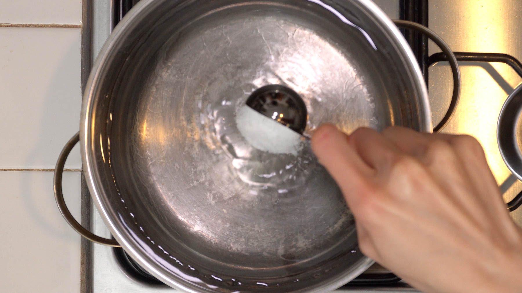 A hand adds salt to a stainless steel pot of water on a stove.