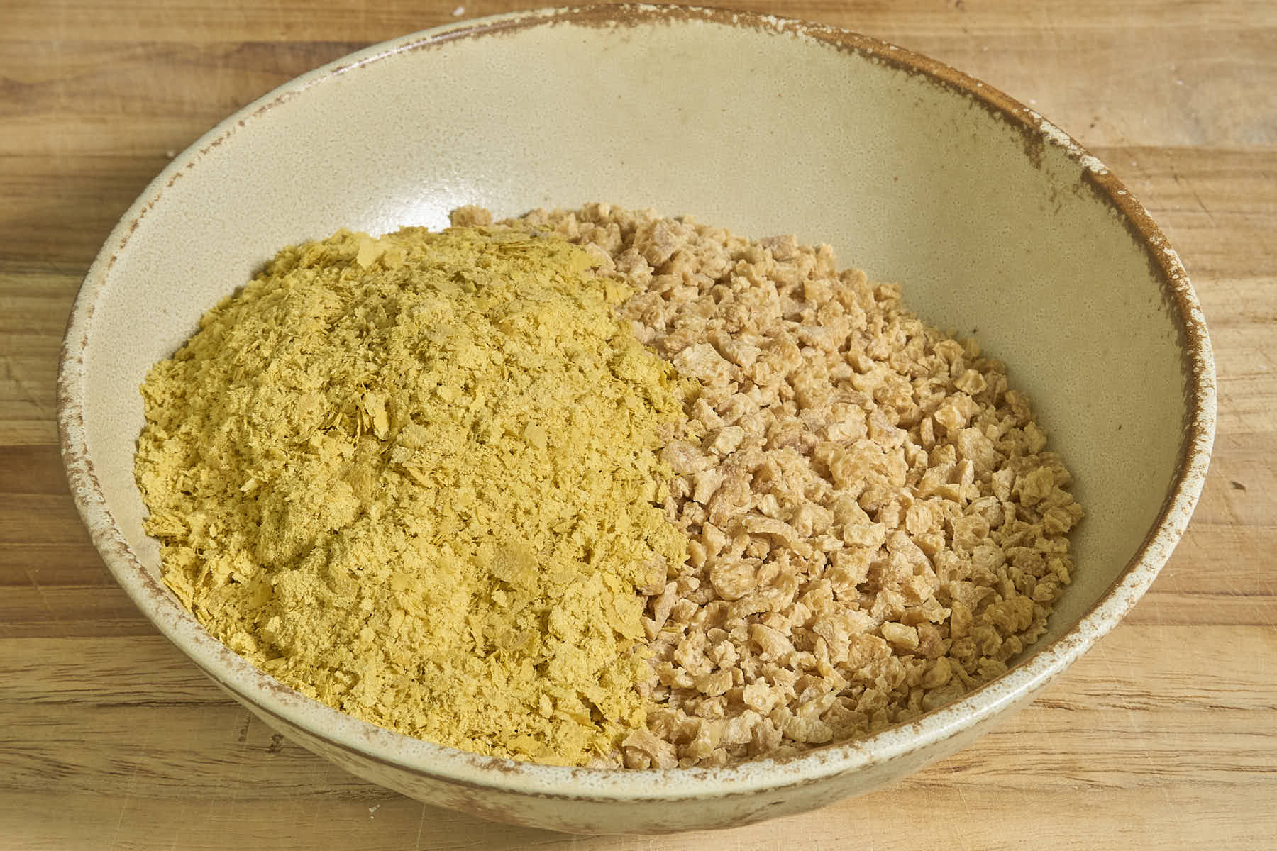 A bowl with yellow nutritional yeast and light brown TVP sits on a wooden surface.